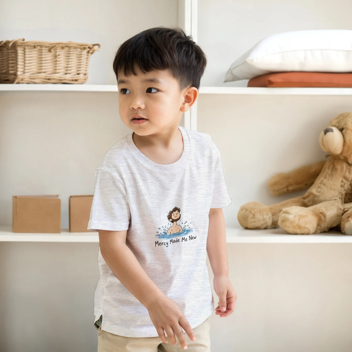 Grey Mercy Made Me New Kids Tee, room mockup with a shelf and teddy bear