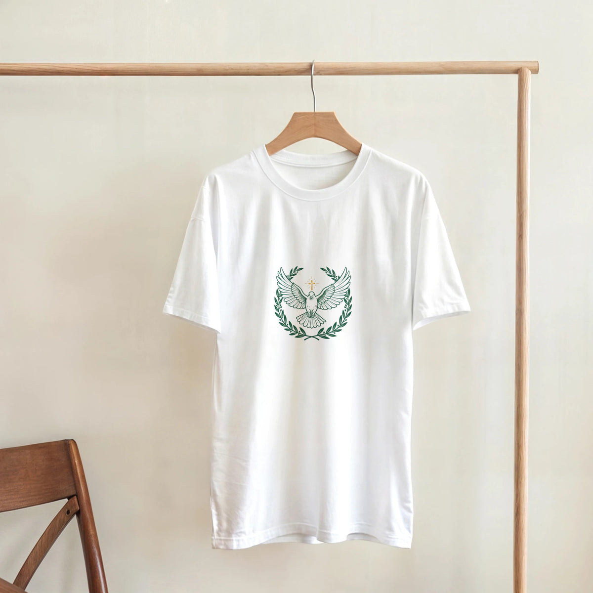 White t-shirt with a dove with wings spread inside an olive branch wreath below a cross, on a wooden hanger